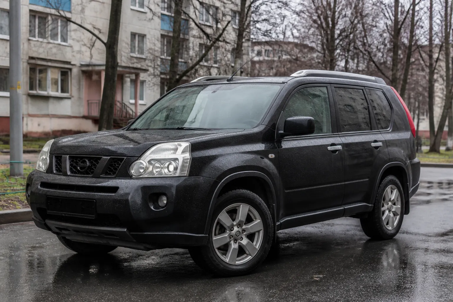 Nissan X-Trail T31 стоит на городской дороге в пасмурную погоду. На фоне видны дома и мокрый асфальт, подчёркивающие реальный сценарий эксплуатации кроссовера.