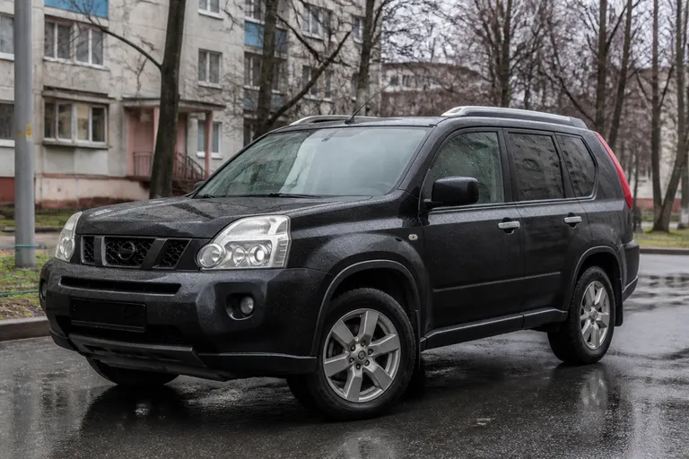 Nissan X-Trail T31 стоит на городской дороге в пасмурную погоду. На фоне видны дома и мокрый асфальт, подчёркивающие реальный сценарий эксплуатации кроссовера.