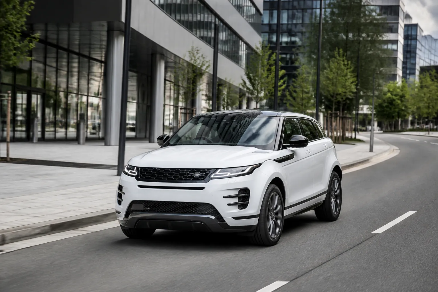 Белый Land Rover Range Rover Evoque на городской дороге у современного здания. Автомобиль снят спереди под углом в дневном естественном свете.