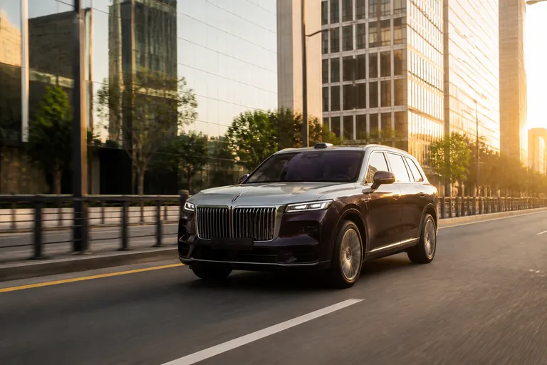 Новый крупный SUV Voyah Taishan на городской дороге в вечернем свете. Автомобиль показан спереди под углом на фоне современного квартала.