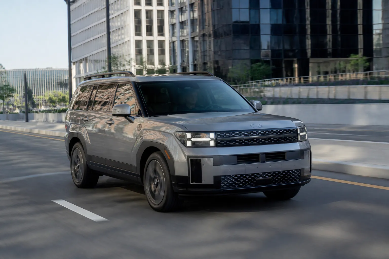 Hyundai Santa Fe нового поколения на городской дороге, крупный семейный кроссовер снят спереди под углом. На фоне современный город и мягкий дневной свет.