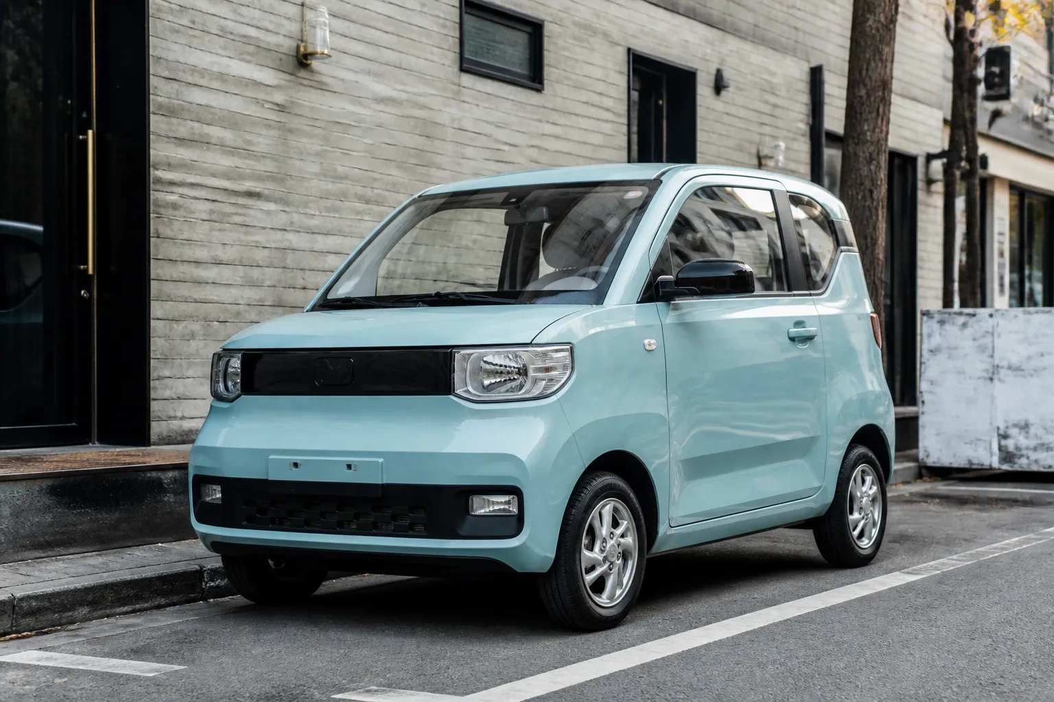 Компактный Wuling Hongguang Mini EV стоит на городской улице рядом с современными зданиями. Машина показана как небольшой электромобиль для повседневных поездок по городу.