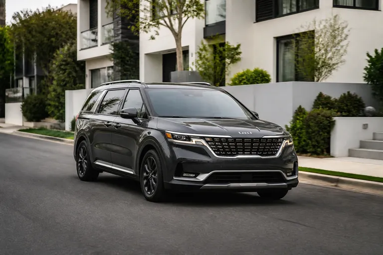 Kia Carnival на городской дороге рядом с семейным домом, автомобиль снят спереди под углом. На фоне видны спокойная улица и мягкий дневной свет.