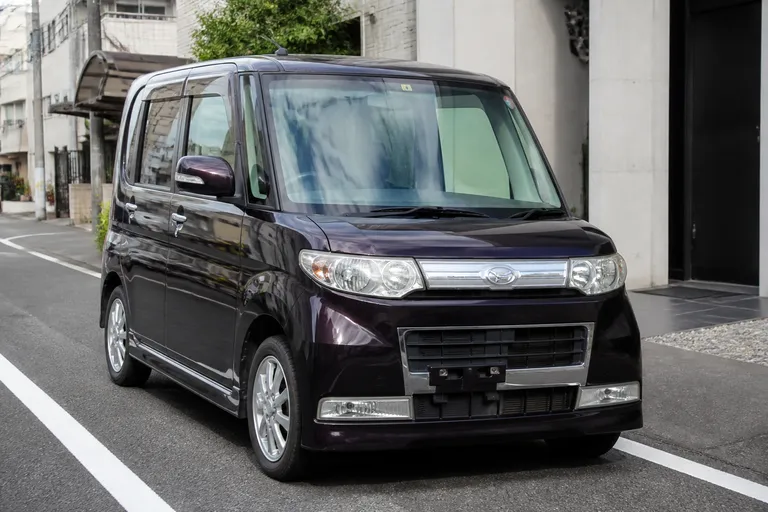 Daihatsu Tanto на городской улице в Японии рядом с разметкой и невысокими зданиями. Автомобиль показан спереди сбоку в естественном дневном свете.