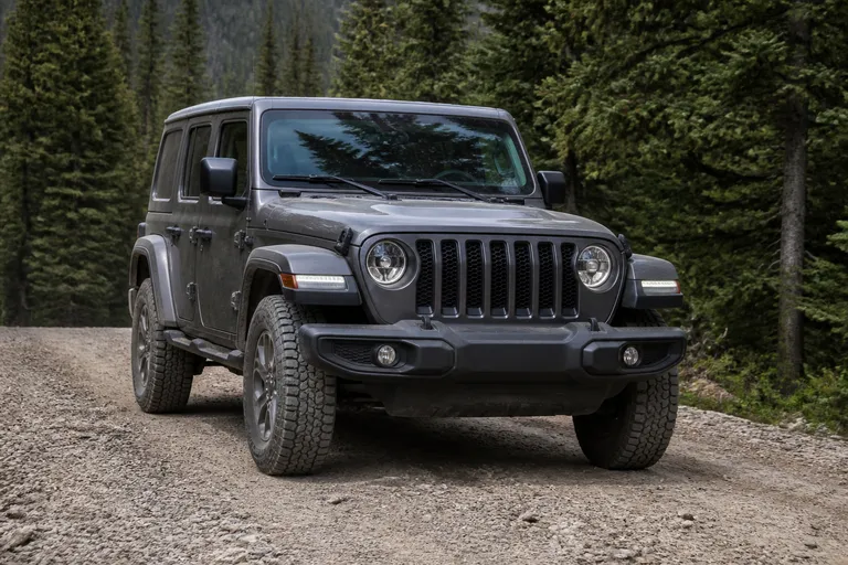 Jeep Wrangler на грунтовой дороге за городом, автомобиль показан спереди под углом. На фоне видны лес и дневной естественный свет.