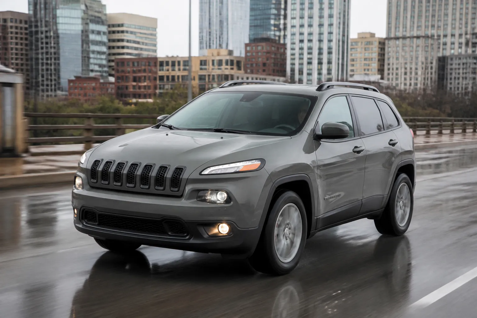 Jeep Cherokee на городской дороге в пасмурный день, снятый спереди под углом. На фоне видны мокрый асфальт и современная городская застройка.