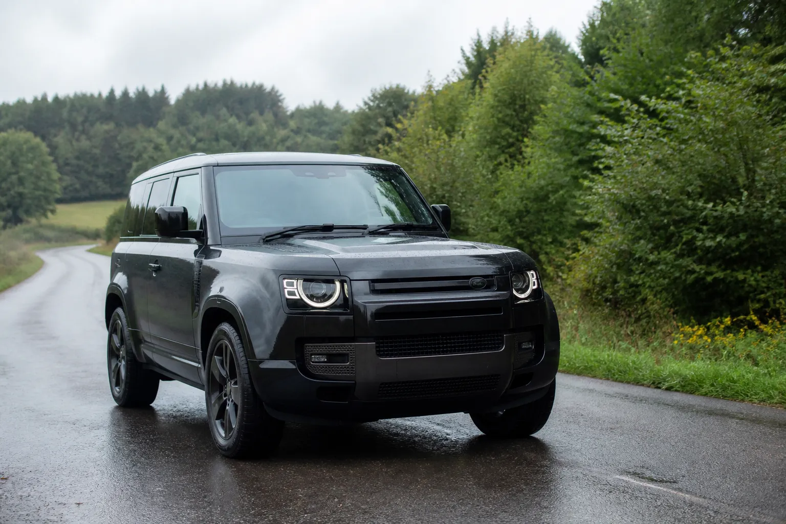 Land Rover Defender на загородной дороге в пасмурную погоду. Автомобиль показан спереди под углом, акцент на кузове и дорожной обстановке.