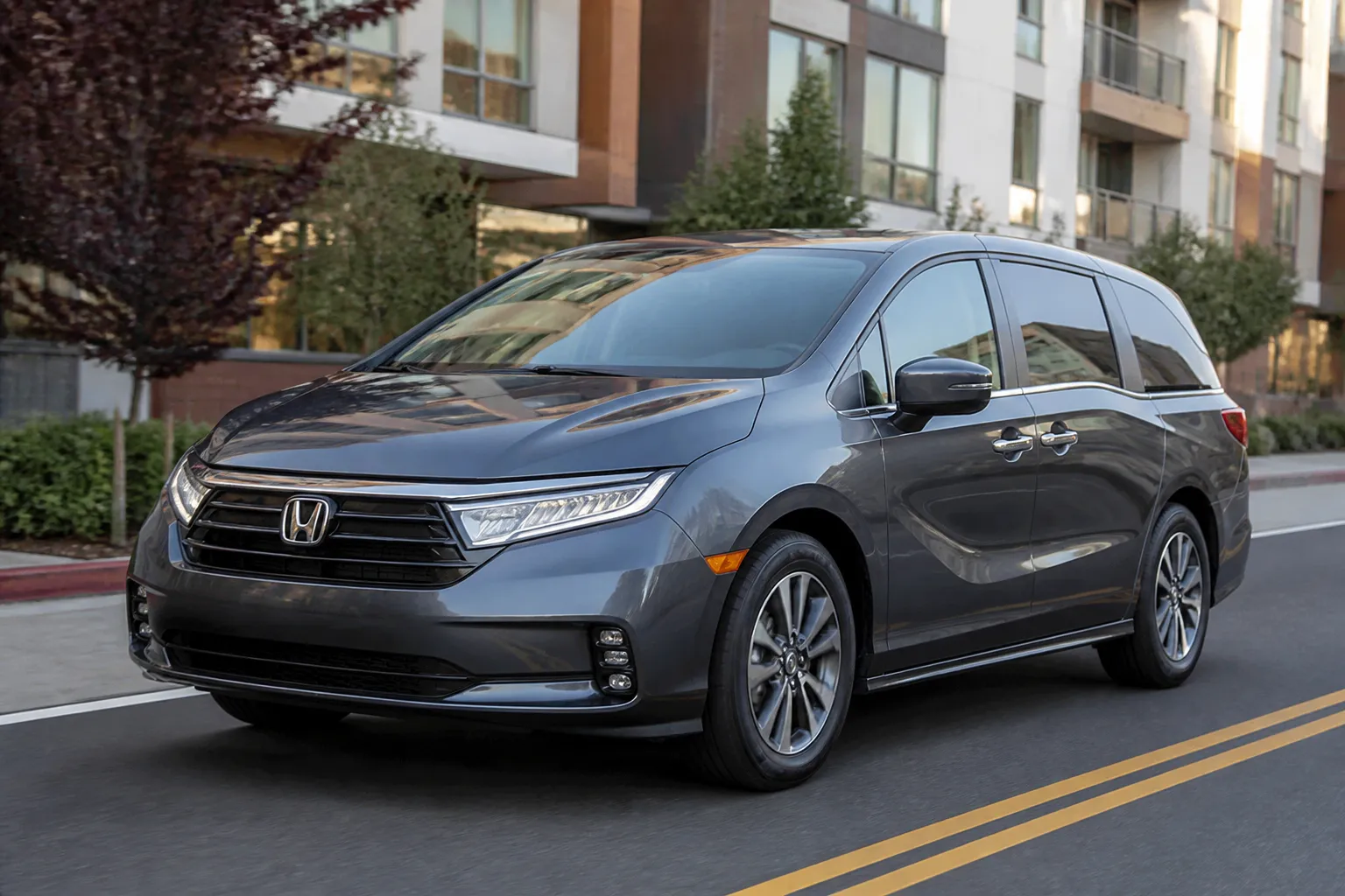 Honda Odyssey на городской дороге у современного жилого квартала. Минивэн показан в естественном свете, с акцентом на семейный формат и практичность.