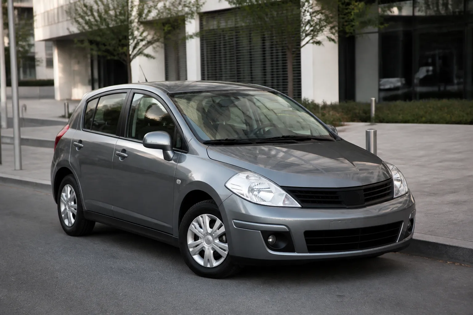 Nissan Tiida на городской улице перед осмотром перед покупкой. Рядом видны дорога, современные здания и нейтральное дневное освещение.